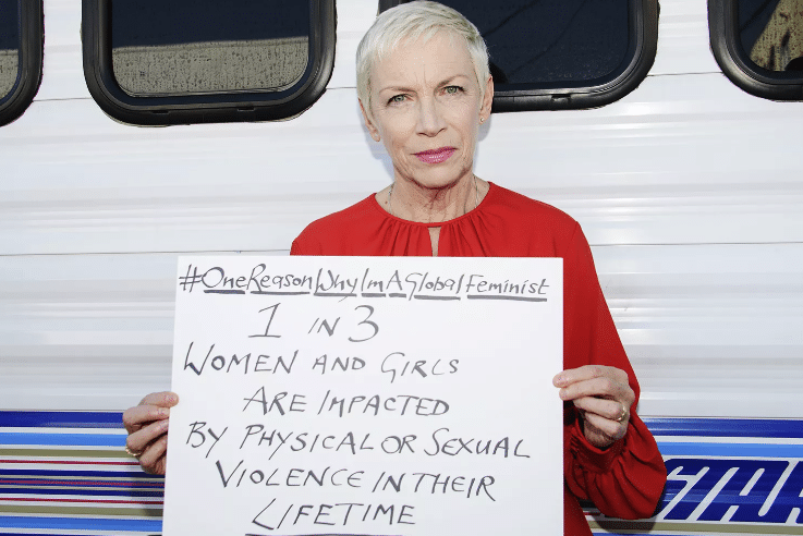 A picture of Annie Lennox, the singer. She is wearing a red flowy shirt and holding a sign that is handwritten in capital letters. It says #one reason why I am a global feminist, underneath this writing reads. 1 in 3 women and girls are impacted by physical or sexual violence in their lifetime. 
