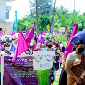 Women on a march for garment justice. Most are carrying pink flags and holding signs. One women is holding an umbrella. 