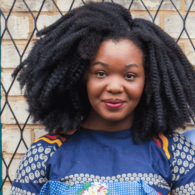 A photo of Amarchi Clarke: she is wearing a colourful blouse with red and yellow stripes and mostly blue tones. She is wearing dark pink lipstick, her hair is styled in an afro and she looks smiley but serious. She is standing behind  a brick wall. 