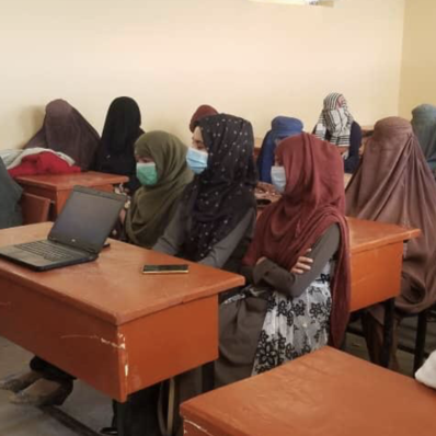 A group of Afghan school girls all wearing masks and hijabs, they are sat at light brown desks and there is a teacher at the front wearing a turqoise dress. The walls of the room are beige and empty. There is a laptop in shot.