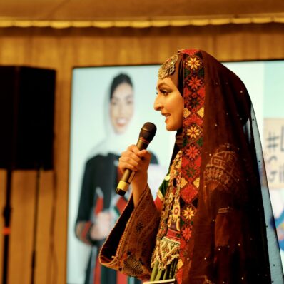 A women is wearing an ornamental red, yellow and black dress with a headscarf and forehead decoration. She is on s stage with a mic and the photo details her in portrait. Behind her on the left is a speaker and on the right a poster that says 