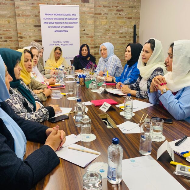 A group of women sat round a table, they are all wearing hijabs of different bright colours and there's scattered paper and water bottles on the table
