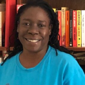 A photo of Dr Atuki Turner, a Uganda scholar, she is wearing a blue t shirt and is smiling. Her backdrop is a book case with red, yellow and orange books